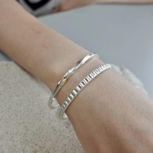 May include: Two silver bangles on a wrist. The top bangle has a twisted design, while the bottom one features a textured, ridged pattern. The jewelry is displayed on a person's arm, showcasing the bracelets' shine.