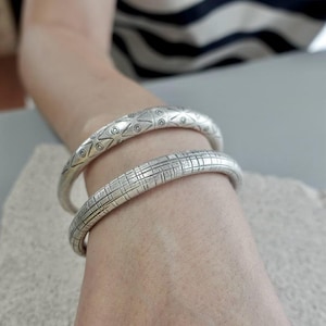 May include: Two silver bangles are worn on a person's wrist. The top bangle has a repeating pattern of circles and triangles. The bottom bangle has a grid-like pattern. The bangles are slightly curved and have a matte finish.