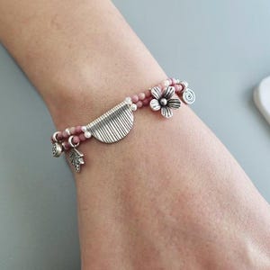 May include: A bracelet with pink and white beads. It features silver-toned charms, including a flower, a leaf, and a half-circle design. The bracelet is worn on a wrist.