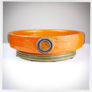 May include: Orange ceramic bowl with a black and white ring design. The bowl has a green and brown base.