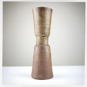 May include: A tall, cylindrical ceramic vase with a distinct hourglass silhouette. The surface features a textured, speckled finish in earthy tones of brown and tan, with a wider base that tapers inward before widening again at the top rim.