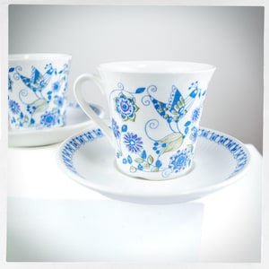 May include: Two white teacups with blue and green floral designs. The cups have a delicate floral pattern with birds and leaves. The cups are on white saucers with a blue and white border.