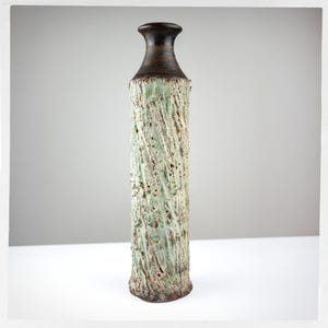 May include: A tall, cylindrical ceramic vase with a narrow neck and a dark brown, smooth finish at the top. The body features a textured, light green and off-white surface with a diagonal, spiraling pattern that resembles rough, weathered bark.
