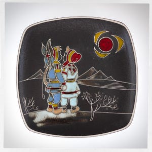 May include: A black ceramic plate featuring a stylised image of two figures in traditional clothing standing in a snowy landscape. The figures are looking up at a red sun with yellow rays. The plate has a textured surface.