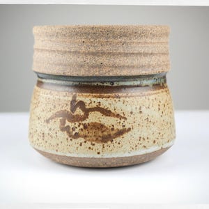 May include: A ceramic cup with a speckled beige and brown glaze. The upper rim features a textured, sandy-colored band, while the body displays a rounded shape with a dark brown hand-painted abstract mark and scattered brown speckles throughout.