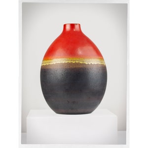 Peut inclure: Un vase décoratif en céramique de forme arrondie. Le haut est rouge, avec une bande dorée, et une base marron foncé. Le vase est présenté sur un socle blanc.