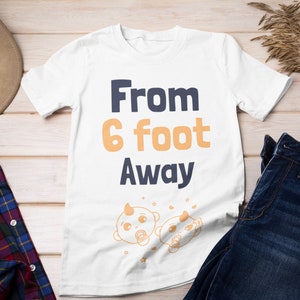 Könnte beinhalten: Weißes T-Shirt mit dem Text "From 6 foot Away" in Marineblau und Orange. Zwei Comic-Babys mit Schnullern befinden sich unter dem Text.