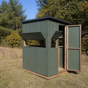 May include: A green hunting blind with an open door and window flaps. The structure is made of green panels with a wooden frame and sits on a wooden base. The background features trees and grass.