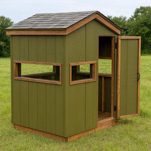 May include: A green wooden hunting blind with a brown shingled roof and wooden trim. The blind has two rectangular windows and an open door, set in a grassy field.