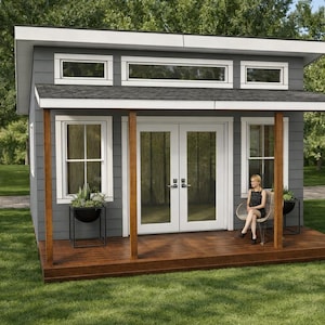 Op de afbeelding: Een kleine, moderne grijze schuur met een bruine houten veranda en witte afwerking. De schuur heeft een dubbele deur en ramen. Een persoon zit op een stoel op de veranda. Potplanten staan op de veranda en bij de ramen.
