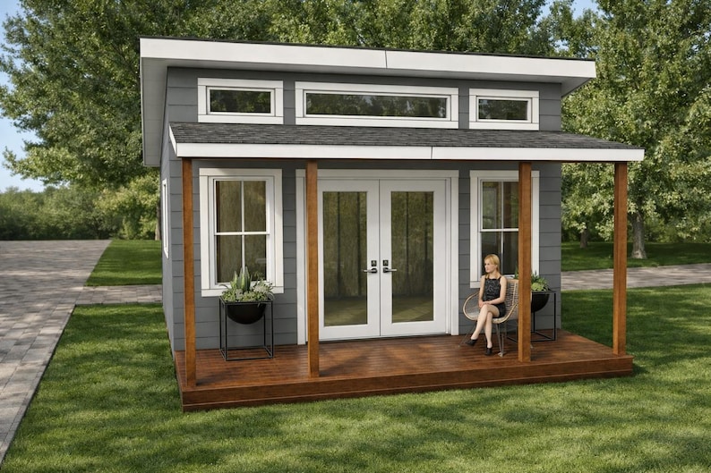 Op de afbeelding: Een kleine, moderne grijze schuur met een bruine houten veranda en witte afwerking. De schuur heeft een dubbele deur en ramen. Een persoon zit op een stoel op de veranda. Potplanten staan op de veranda en bij de ramen.