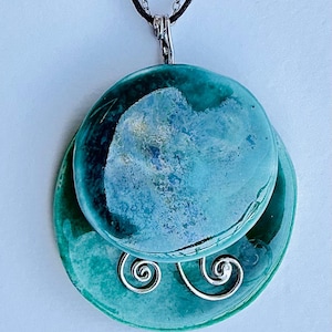 May include: A teal ceramic pendant necklace. The pendant has two circular layers, the top one lighter than the bottom. The top layer has a swirling pattern. Silver wire swirls are attached to the bottom layer. The pendant hangs from a black cord.