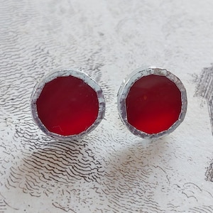 Puede incluir: Un par de pendientes de botón redondos con un centro rojo vibrante y un borde plateado texturizado. Los pendientes se presentan sobre un fondo con un diseño artístico vintage.