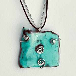 May include: A turquoise pendant necklace with an irregular, rectangular shape. The pendant has a glossy finish and is decorated with silver wire swirls. The necklace has a black cord and a silver chain.