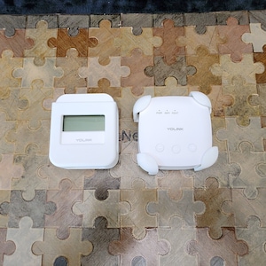May include: Two white electronic devices. One is rectangular with a screen, the other is square with rounded edges. The square device has the text "PWR WIFI FEAT" and the brand name "YOLINK" is visible on both devices.