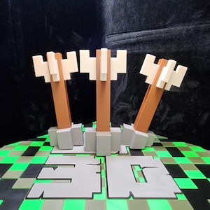 May include: A 3D-printed sculpture featuring three stylized axes with brown handles and white axe heads. The axes are mounted on a gray base with the word "3D" in gray block letters. The base sits on a green and black checkered surface.