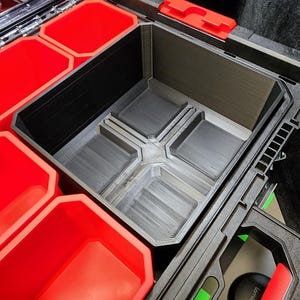 Puede incluir: Un organizador de plástico negro y gris con compartimentos rojos. El organizador tiene un compartimento central grande y varias secciones más pequeñas. Los compartimentos están diseñados para guardar herramientas u otros objetos pequeños. El organizador está abierto y listo para usar.