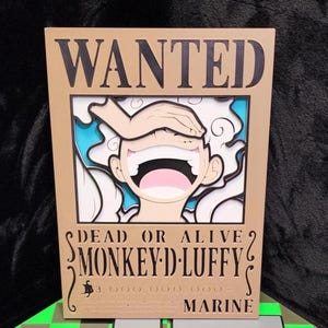 May include: A rectangular tan sign featuring the word WANTED in black at the top. Below, a layered illustration shows a laughing character with a hand over their eyes. Text reads DEAD OR ALIVE, MONKEY D. LUFFY, 3,000,000,000, and MARINE in black.