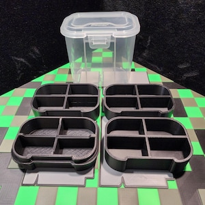 May include: A clear plastic storage container with a white lid and four black, divided trays. The trays are arranged on a green and black checkered surface, suggesting an organizational theme.