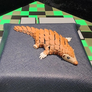 Può includere: Scultura di pangolino in legno con corpo segmentato e strutturato nei toni del marrone e beige. Il pangolino ha zampe bianche e un muso chiaro. Poggia su una superficie blu scuro strutturata.