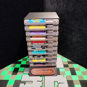 Puede incluir: Una pila de cartuchos de videojuegos clásicos de Nintendo, que incluyen títulos como "Tetris" y "Double Dragon". Los cartuchos grises tienen etiquetas coloridas y se exhiben en un soporte negro con el logotipo de Nintendo. La base está sobre una superficie a cuadros verdes y grises.