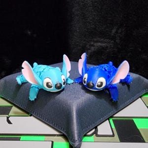 Puede incluir: Dos pequeñas figuras azules y turquesas de un personaje de dibujos animados con orejas grandes y ojos grandes. Las figuras están sobre una superficie azul oscuro con un patrón geométrico. El personaje tiene un cuerpo texturizado y orejas internas rosas.