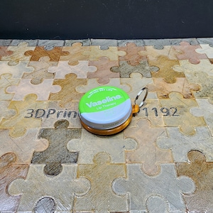 May include: A small, round, green and white tin of Vaseline Lip Therapy with a gold lid and a silver key ring attached. The tin is sitting on a brown and orange puzzle mat.