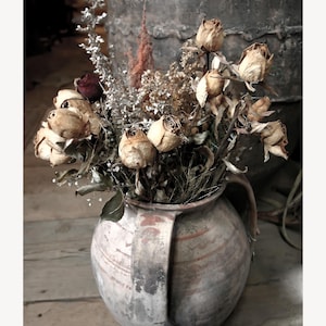 May include: A rustic, aged ceramic vase with a handle, filled with dried roses and other dried flowers. The vase is a muted brown and gray, and the flowers are a mix of beige, brown, and red. The arrangement sits on a wooden surface.