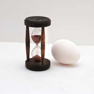 May include: A vintage hourglass with dark wood accents and a clear glass chamber filled with reddish-brown sand. Beside it is a white egg, creating a contrast in textures and colors. The hourglass is approximately 4 inches tall.