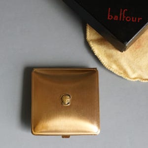 May include: A gold-colored metal compact with a raised crest in the center. The compact is square and has a hinged lid.