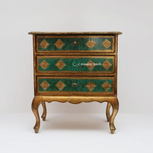 May include: A small, antique-style chest of drawers with three drawers. The drawers are teal with gold filigree designs. The frame and legs are gold-colored with a distressed finish. The chest has a missing knob on the middle drawer.