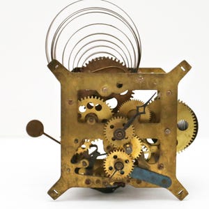 May include: A close-up of a brass clock mechanism with a coiled spring and intricate gears. The clock face is missing, but the hands are visible.