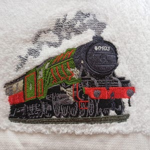 May include: A white terry cloth towel featuring an embroidered steam locomotive in black, green, and red thread. The train design includes the number 60103 on the front and grey embroidered smoke billowing from the top of the engine.