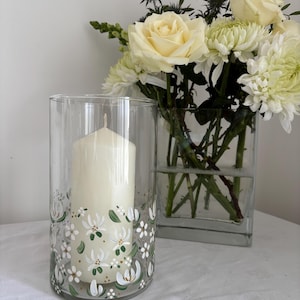 May include: A clear glass candle holder with a white pillar candle inside. The holder is decorated with a floral design in white, green, and gold. A square glass vase with white roses and chrysanthemums is in the background.