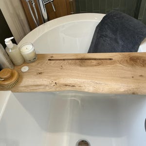 Puede incluir: Una bandeja de baño de madera con un acabado de madera natural. La bandeja tiene una ranura en el centro para sujetar un libro o una tableta. Hay dos botellas de productos de baño, una vela y un cepillo de baño en la bandeja.
