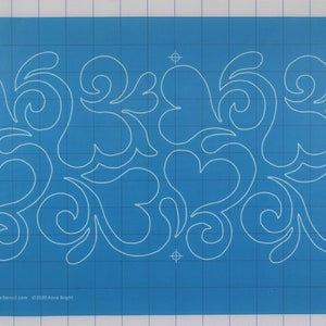 May include: A blue quilting template with a white design of swirling hearts and leaves. The template is on a gridded surface. The text "#00527 www.fullcircletemplates.com ©2023 Anne Bright" is in the bottom left corner.