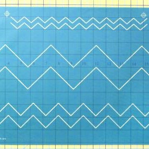 May include: A blue plastic stencil with three rows of white zig-zag patterns. The stencil is marked with grid lines and numbers.
