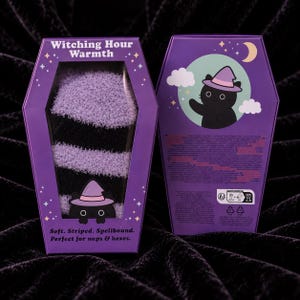 May include: A pair of fuzzy black and lavender striped socks packaged in a coffin-shaped purple box. The box features the text Witching Hour Warmth and Soft. Striped. Spellbound. Perfect for naps & hexes. A black cat wearing a purple hat is illustrated.