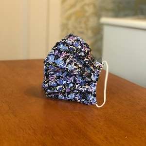 Op de afbeelding: Een blauw en paars bloemenpatroon mondkapje met witte elastische oorlussen.