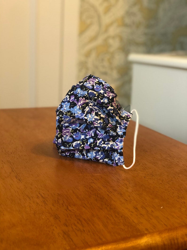 Op de afbeelding: Een blauw en paars bloemenpatroon mondkapje met witte elastische oorlussen.
