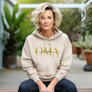 Könnte beinhalten: Ein beiger Hoodie mit einem goldenen Aufdruck "OMA Karl EST. 2023".
