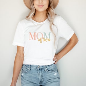 Könnte beinhalten: Weißes T-Shirt mit dem Text "MOM of two" in rosa, blau und goldenen Buchstaben.