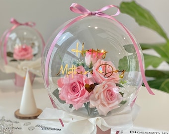 Peut inclure: Un ballon transparent rempli de roses roses et de verdure, avec un ruban rose noué autour de la base. Le ballon est posé dans un pot rose et porte une inscription dorée qui dit "Joyeuse fête des mères".