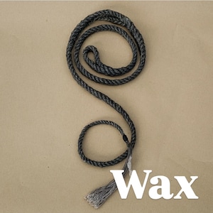 Puede incluir: Una cuerda gris oscura enrollada en forma de espiral, con una borla en el extremo. La palabra "Wax" se muestra en letras blancas en la parte inferior de la imagen. La cuerda está sobre un fondo beige.