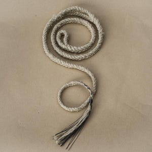 May include: A coiled, off-white rope with a tassel at the end. The rope is thick and appears to be made of natural fibers. The tassel is a mix of off-white and dark gray strands. The rope is displayed on a tan background.