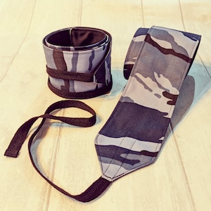 May include: Two weightlifting wrist wraps and a wrist support. The wraps and support feature a blue, grey, and black camouflage pattern. The wrist support has a loop for the thumb. The wrist wraps are rolled up and secured with a black strap.