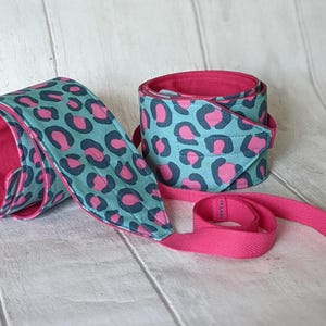 May include: Two weightlifting wrist wraps. The wraps feature a teal leopard print with pink spots and a pink interior. One wrap is rolled up, and the other is unrolled.