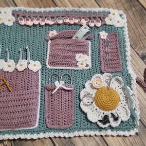 Könnte beinhalten: Ein gehäkelter Nadelhalter mit mehreren Taschen und einem Gänseblümchen-Design. Die Taschen sind mit verschiedenen Näh- und Strickutensilien gefüllt, darunter Nadeln, Scheren und Garn.