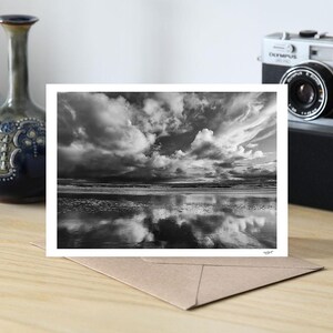 May include: A monochrome photograph of a dramatic cloudscape reflected in water, presented as a greeting card. The card is on a brown envelope, with a vintage camera and decorative vases in the background.