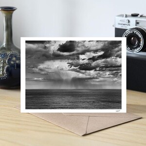 May include: Monochrome photograph of a seascape with a stormy sky. The image is on a white card, resting on a brown envelope. Decorative vases and a vintage camera are in the background.